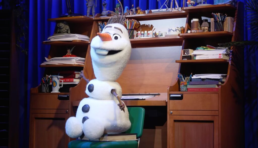 Olaf aparece diante de uma mesa de animação em uma apresentação que aproxima o público